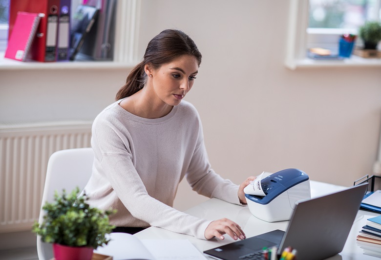 Femeia stătea uitându-se la laptop imprimând o etichetă de pe un dispozitiv din stânga ei.