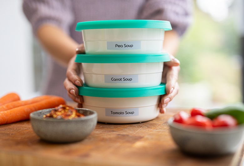 Femeie ținând trei cutii Tupperware etichetate pe o masă de tocat pe care are morcovi, roșii și castraveți