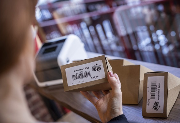 O femeie care verifică etichetele tipărite de un aparat de etichetat Brother, ale alimentelor și care au fost adăugate la sandvișurile preambalate într-un magazin de produse alimentare sau magazin de panificație. Etichetele conțin un cod de bare, titlu, imagine, preț și dată de expirare.