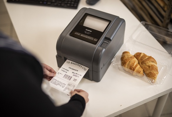 Prim plan cu o imprimantă de etichete Brother imprimând o etichetă pentru alimente, pentru un pachet de cornuri în back office-ul unui magazin de delicatese, al unei brutării sau al unei cafenele